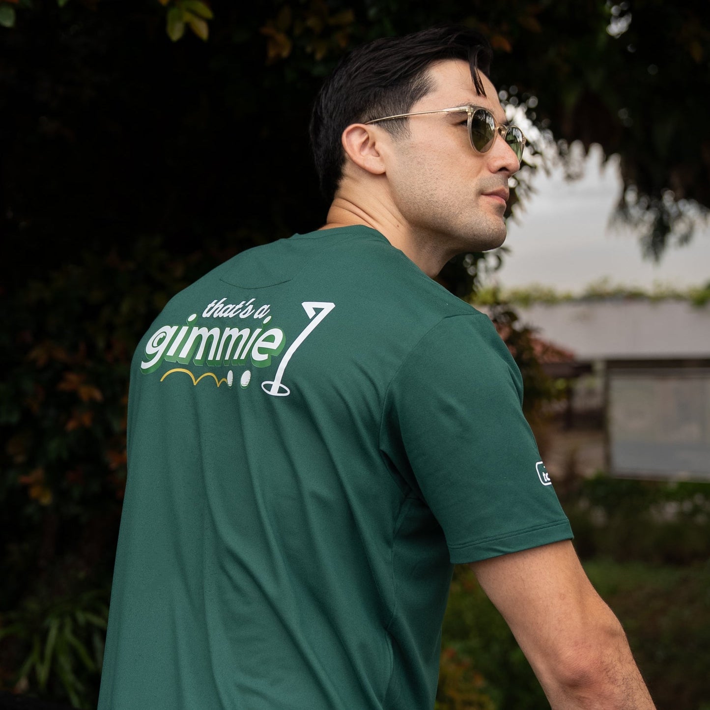 Man wearing a green t-shirt with a brand logo, standing outdoors with trees in the background.