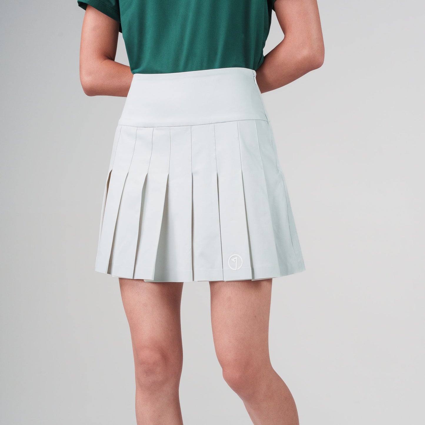 Person wearing a white pleated skirt with a green top and beige sneakers on a light gray background